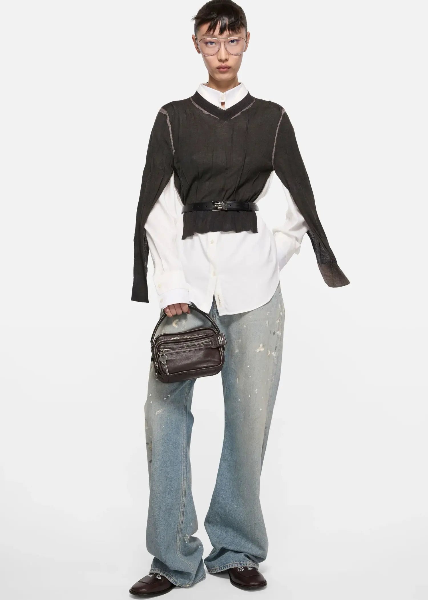 Acne Studios Camero Kit Handtasche in Dunkelbraun mit Trage Riemen und Reißverschluss, Modelfoto von vorne im Studio, Tasche kombiniert mit ausgefallenem Outfit vor weißem Hintergrund
