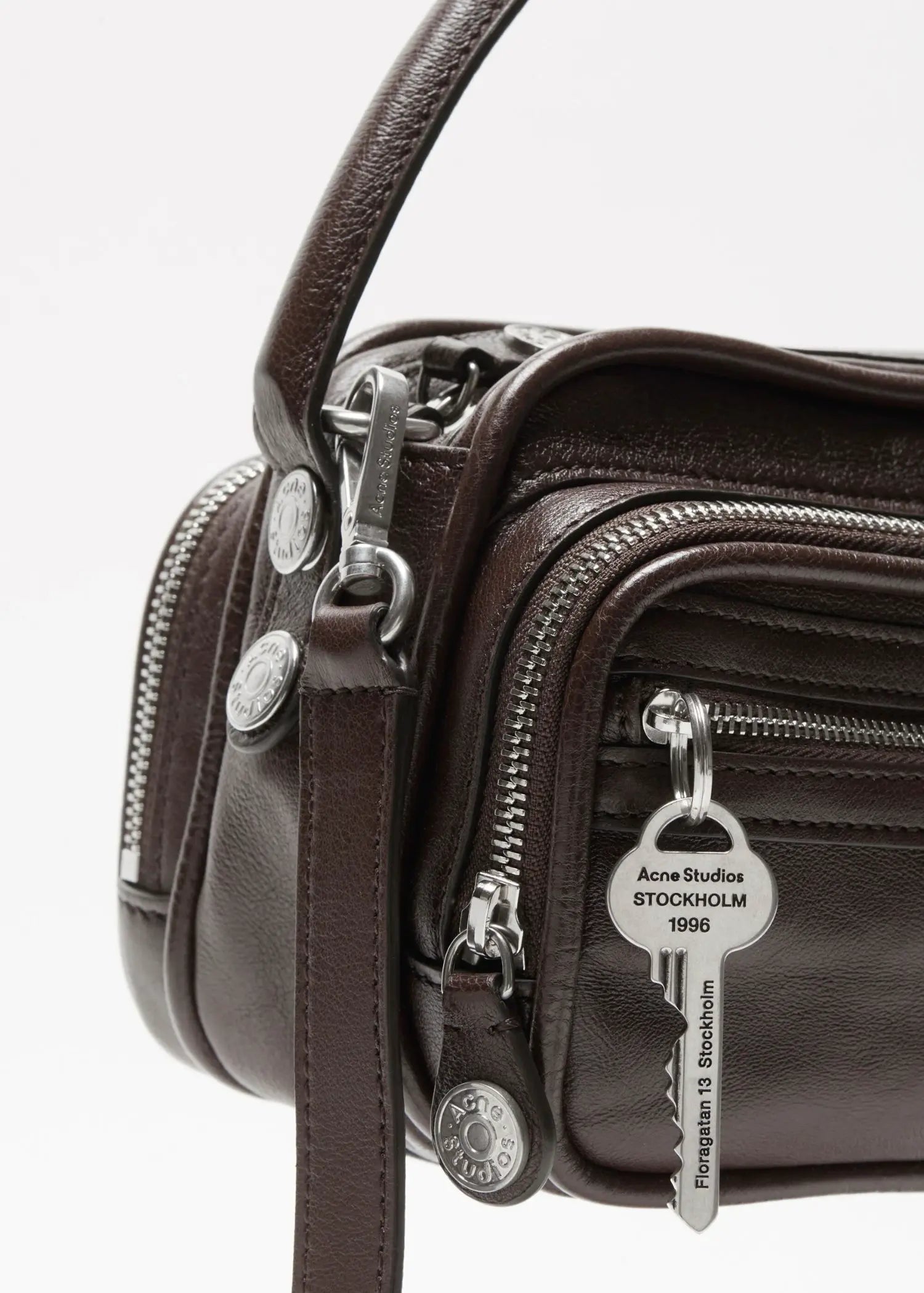 Acne Studios Handtasche aus Kalbsleder, dunkelbraun Nahansicht mit Fokus auf Reißverschluss, Knöpfe, Anhänger und Trageriemen, Studiofoto vor weißem Hintergrund,Produktbild