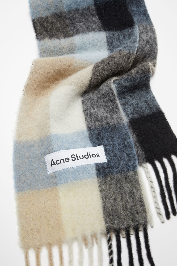 Schal aus Mohair Mischung, Kariert in einer hellen Farbmischung als Creme/Taupe Farben sowie Blau Tönen. Logo Etikett in Weiß. Detail Shot Logo.