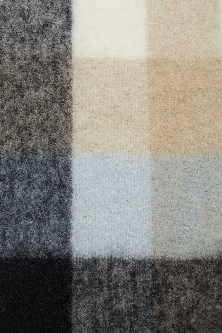 Schal aus Mohair Mischung, Kariert in einer hellen Farbmischung als Creme/Taupe Farben sowie Blau Tönen. Logo Etikett in Weiß. Detail Shot Musterung.