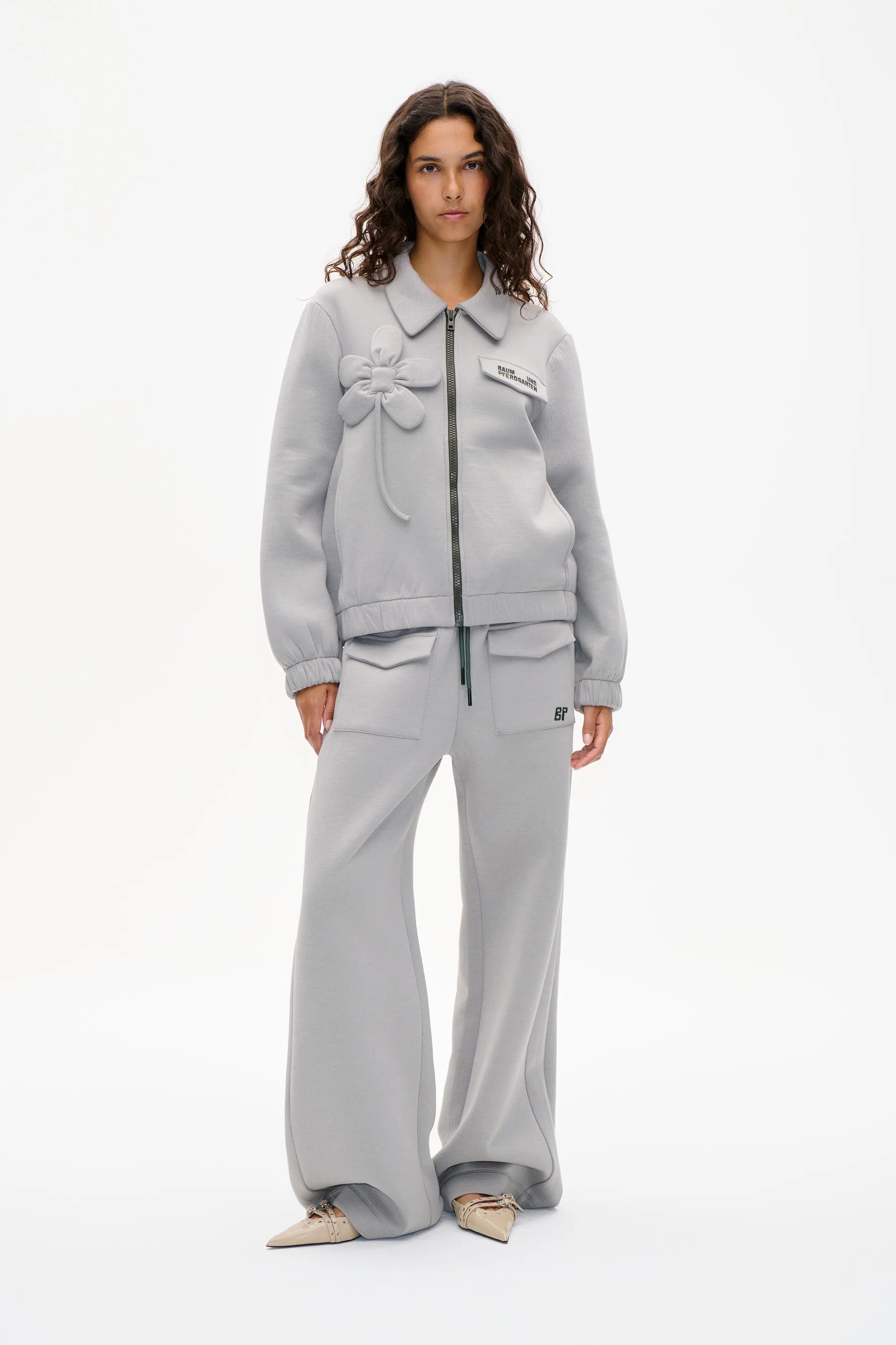 Graue Zipper Jacke, mit aufgenähtem Blumendetail. Modelbild.