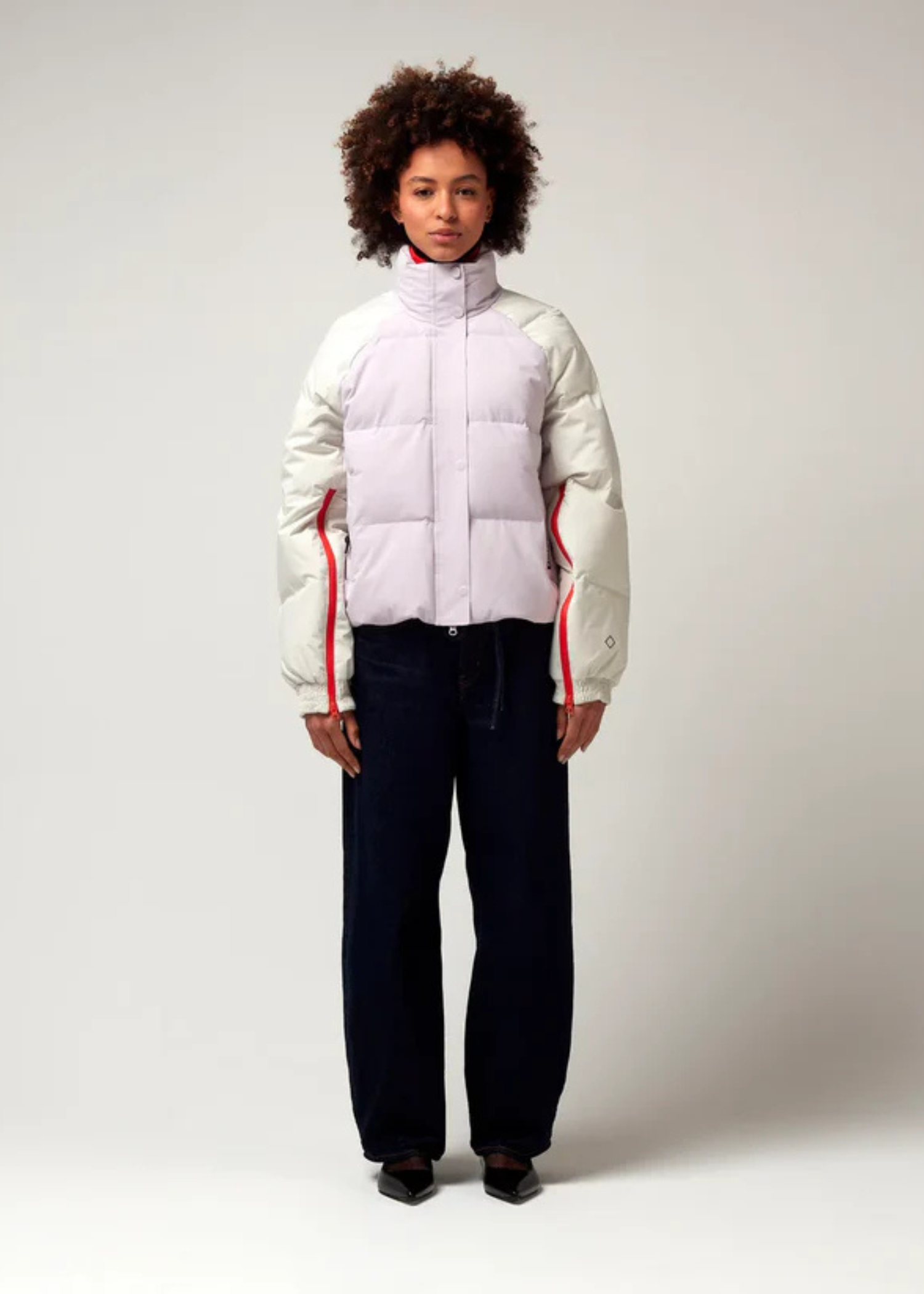 Kurz geschnittene Jacke in Creme, Rosa mit Roten Details. Die Jacke verfügt über einen Stehkragen, einen Reißverschluss mit magnetischer Knopfleiste sowie Reißverschlüssen an den Ärmel, hochreichend bis zur Armbeuge. Model Bild.