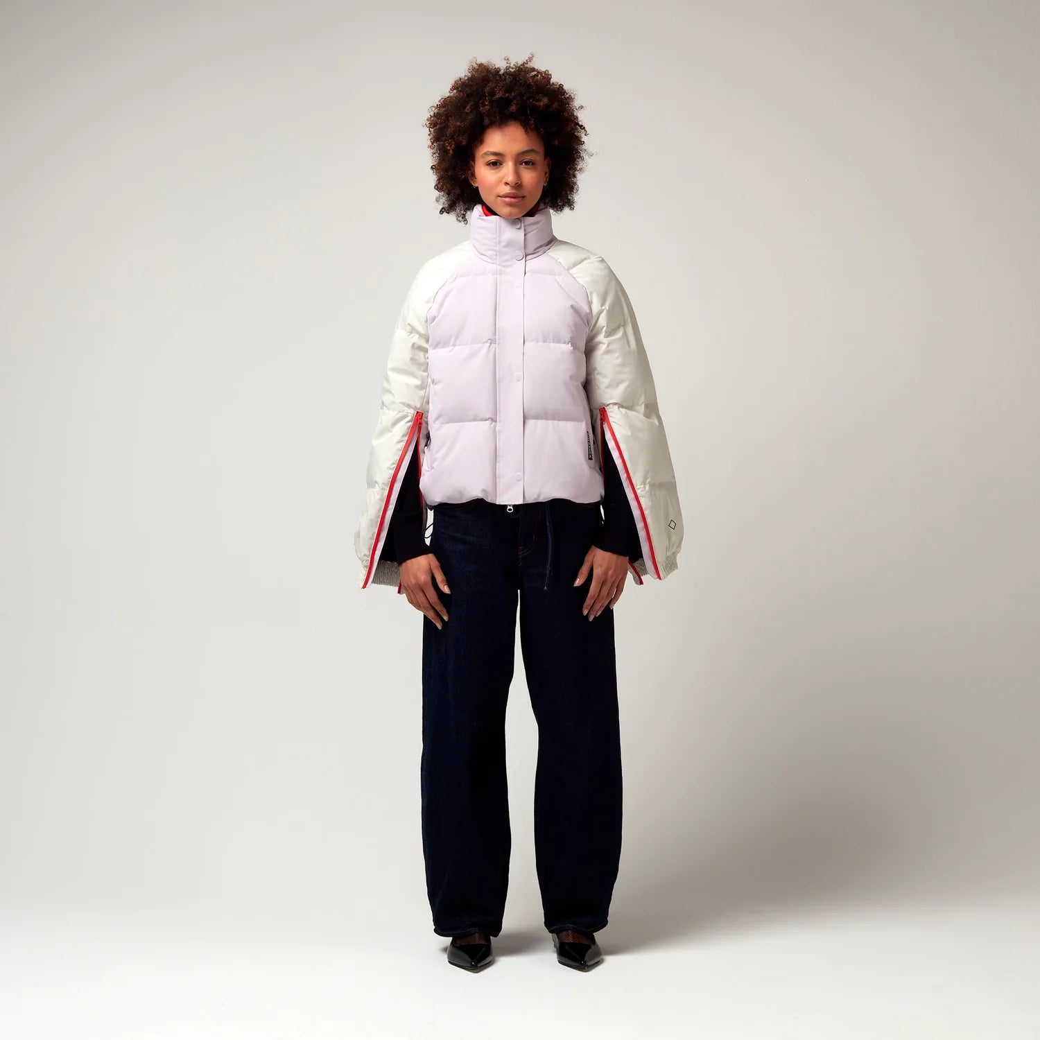 Kurz geschnittene Jacke in Creme, Rosa mit Roten Details. Die Jacke verfügt über einen Stehkragen, einen Reißverschluss mit magnetischer Knopfleiste sowie geöffnete Reißverschlüssen an den Ärmel, hochreichend bis zur Armbeuge. Model Bild.