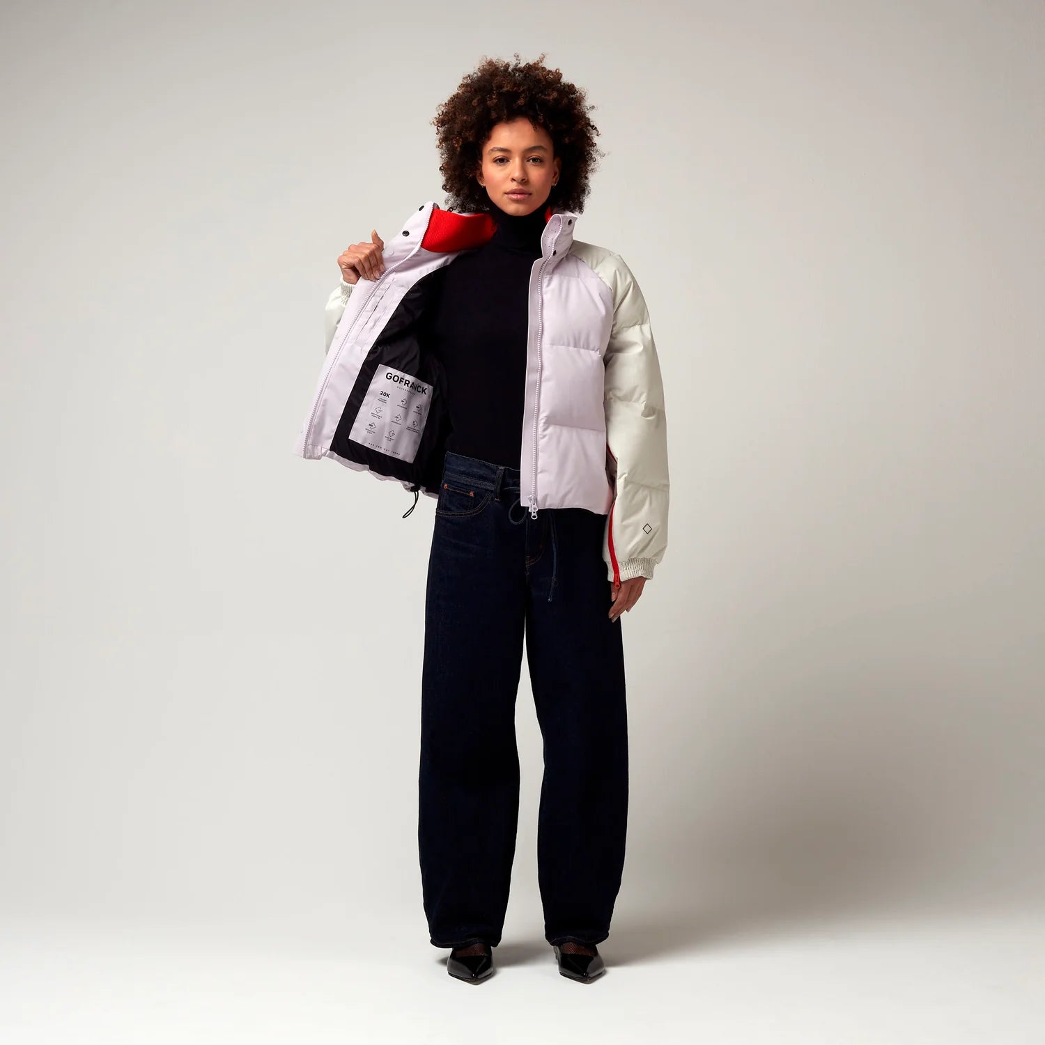 Kurz geschnittene Jacke in Creme, Rosa mit Roten Details. Die Jacke verfügt über einen Stehkragen, einen Reißverschluss mit magnetischer Knopfleiste sowie Reißverschlüssen an den Ärmel, hochreichend bis zur Armbeuge. Leicht geöffnete Jacke, Innenfutter erkennbar. Model Bild.