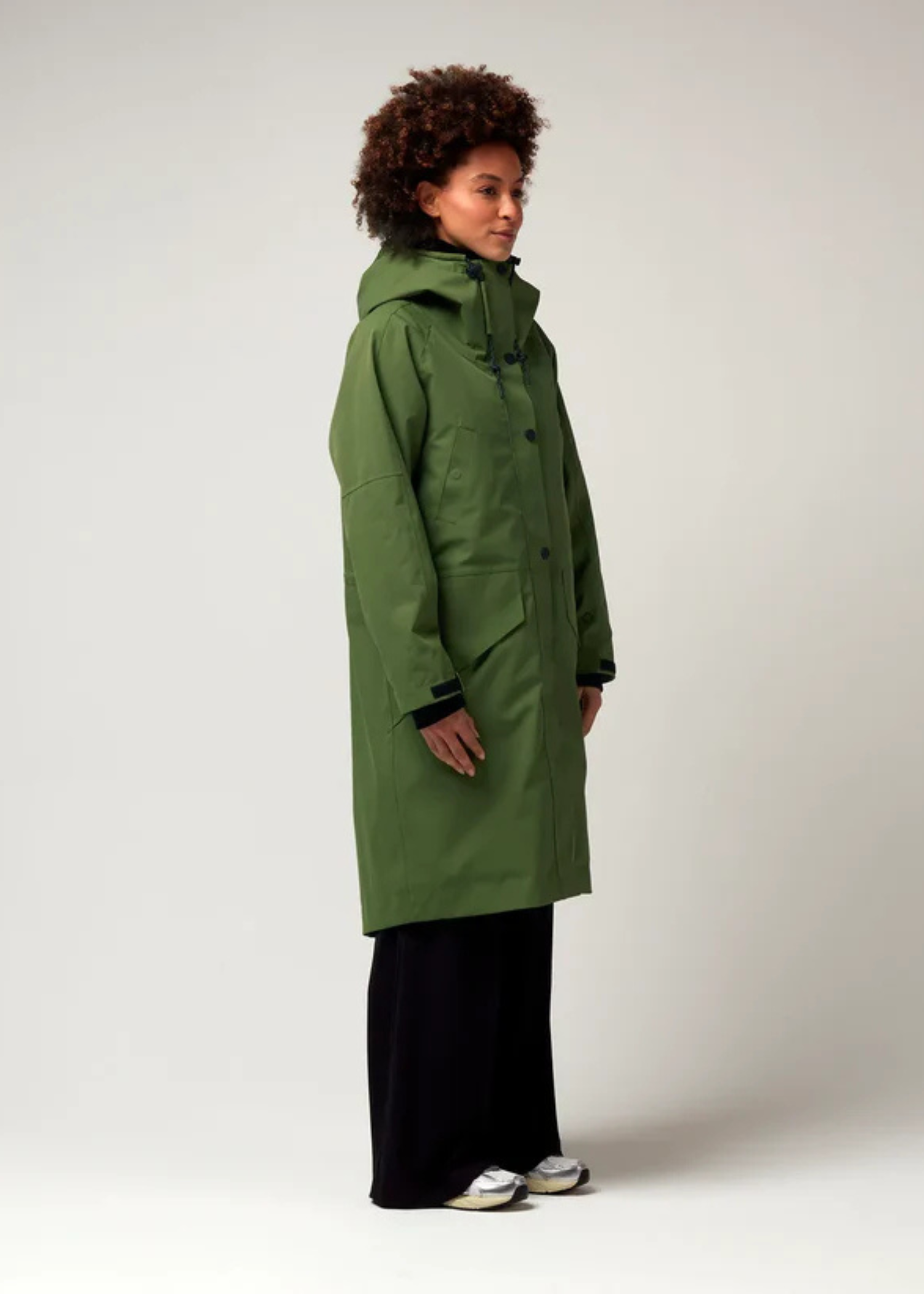 Grüner Parker, mit verstellbarer Kapuze, Seitentaschen sowie Saum mit Daumen Loch. Glatte Oberfläche. Schwarze Hardware. Model Foto Ganzkörper Aufnahme, leicht seitliche Ansicht.