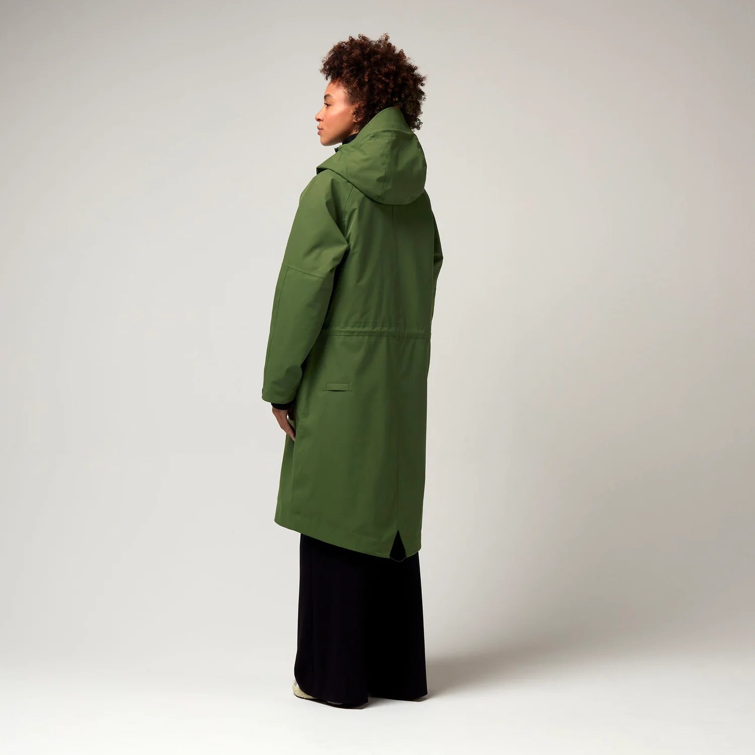 Grüner Parker, mit verstellbarer Kapuze, Seitentaschen sowie Saum mit Daumen Loch. Glatte Oberfläche. Schwarze Hardware. Model Foto Ganzkörper Aufnahme, leicht seitliche Ansicht, Rückseite.