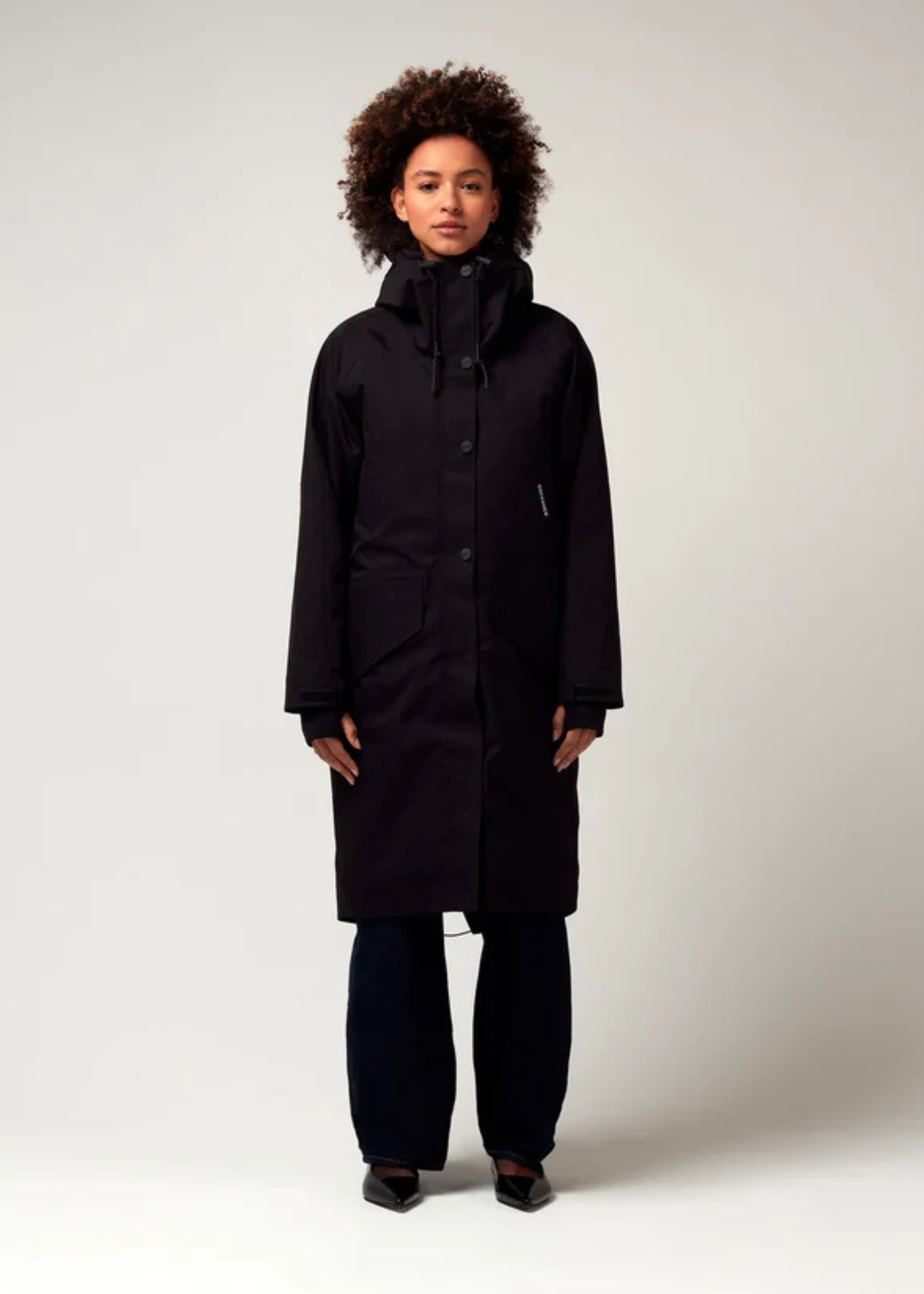 Schwarzer Parker, mit verstellbarer Kapuze, Seitentaschen sowie Saum mit Daumen Loch. Glatte Oberfläche. Model Foto Ganzkörper Aufnahme.