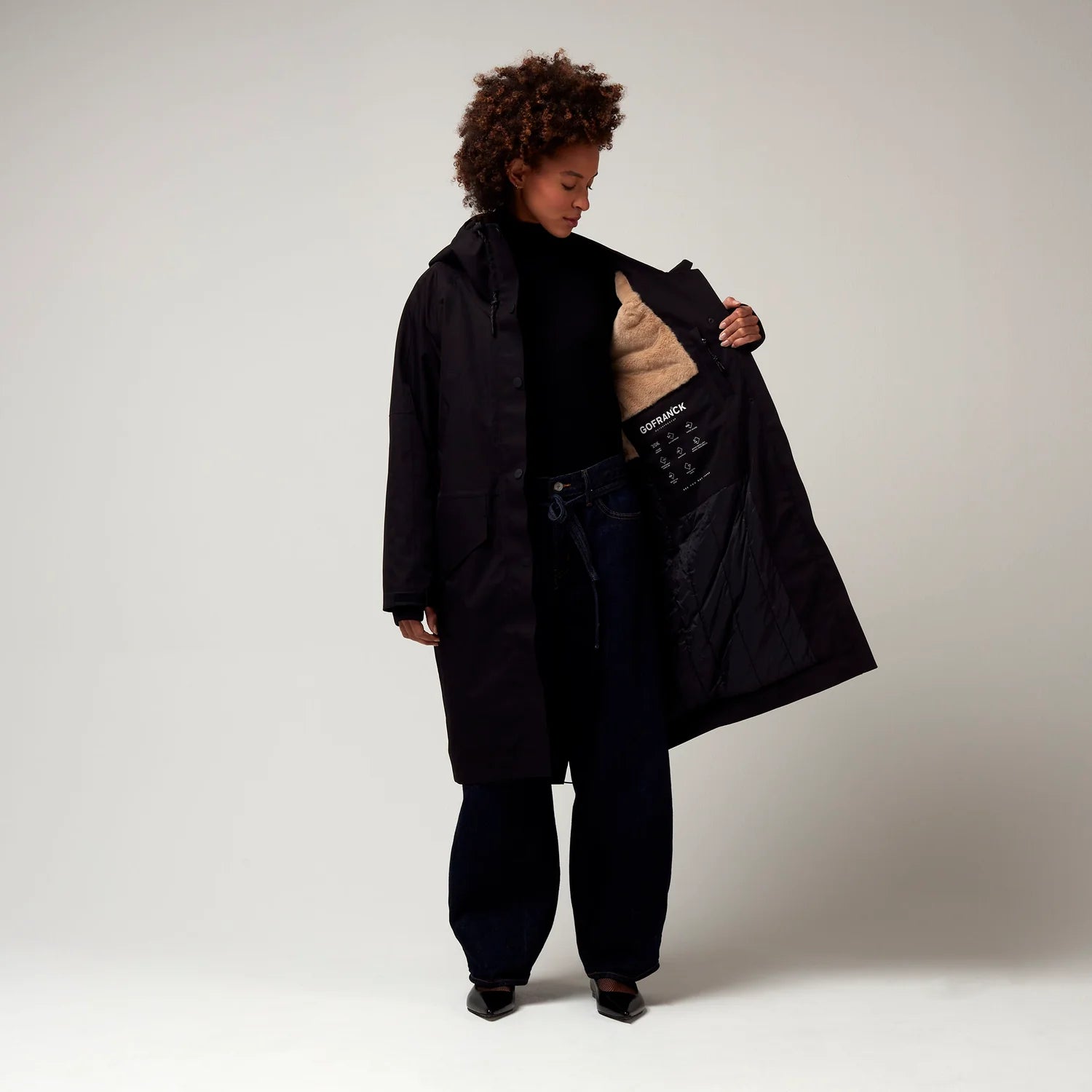 Schwarzer Parker, mit verstellbarer Kapuze, Seitentaschen sowie Saum mit Daumen Loch. Glatte Oberfläche. Model Foto Ganzkörper Aufnahme, leicht geöffnete Jacke mit erkennbarem Innenfutter.