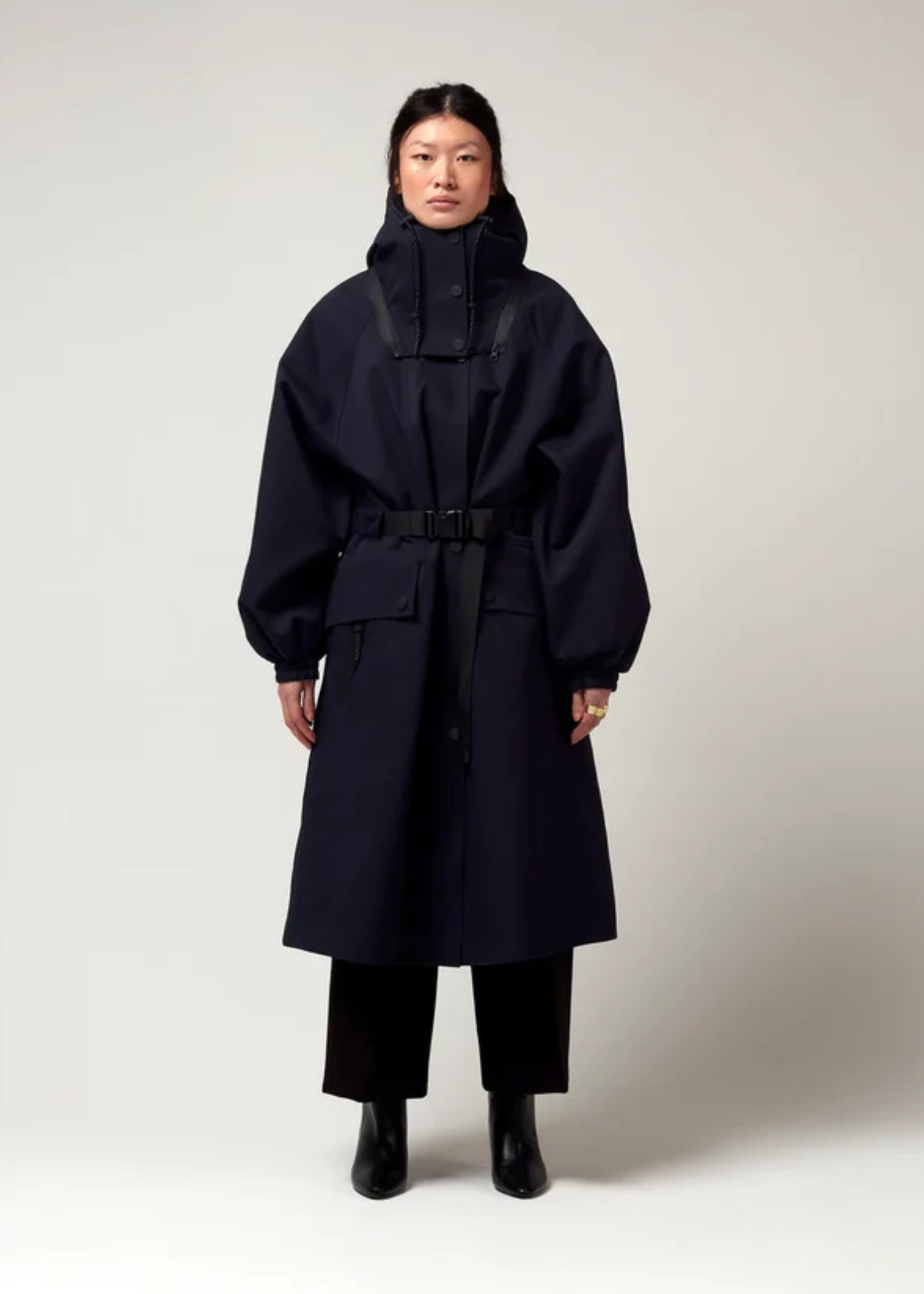 Dunkel Blauer Parker, mit Verstellbarer sowie abnehmbarer Kapuze, Taillierungs- Gurt um die Hüfte und Seiten Taschen. Model Bild, Ganzkörper Aufnahme.