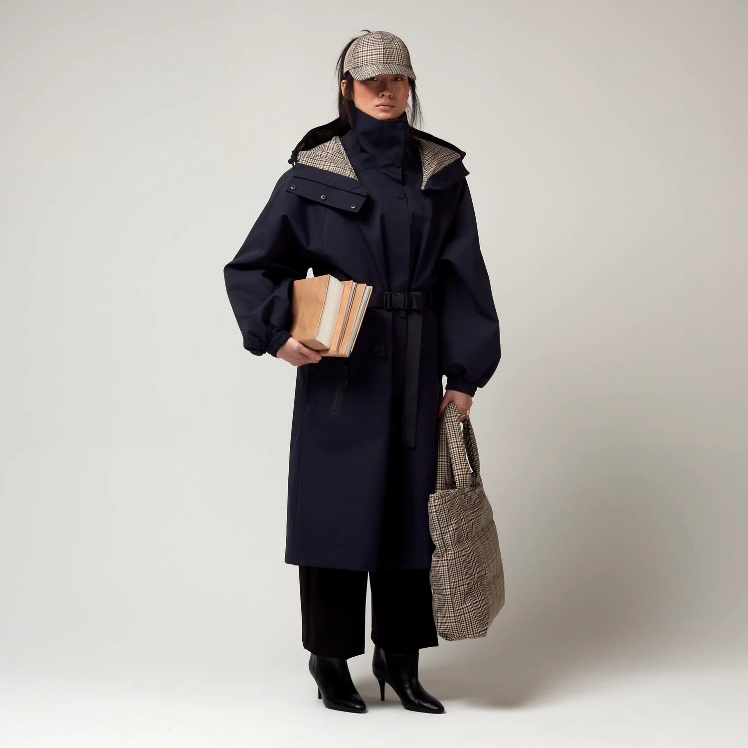 Dunkel Blauer Parker, mit Verstellbarer sowie abnehmbarer Kapuze mit Karo Muster, Taillierungs - Gurt um die Hüfte und Seiten Taschen. Model Bild, Ganzkörper Aufnahme, seitliche Ansicht.