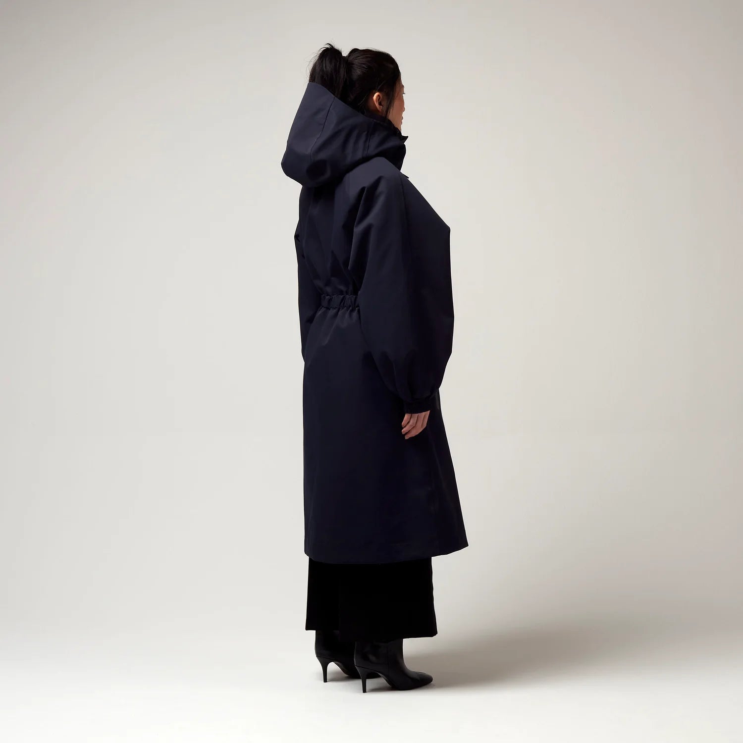 Dunkel Blauer Parker, mit Verstellbarer sowie abnehmbarer Kapuze, Taillierungs - Gurt um die Hüfte und Seiten Taschen. Model Bild, Ganzkörper Aufnahme, seitliche sowie hintere Ansicht.