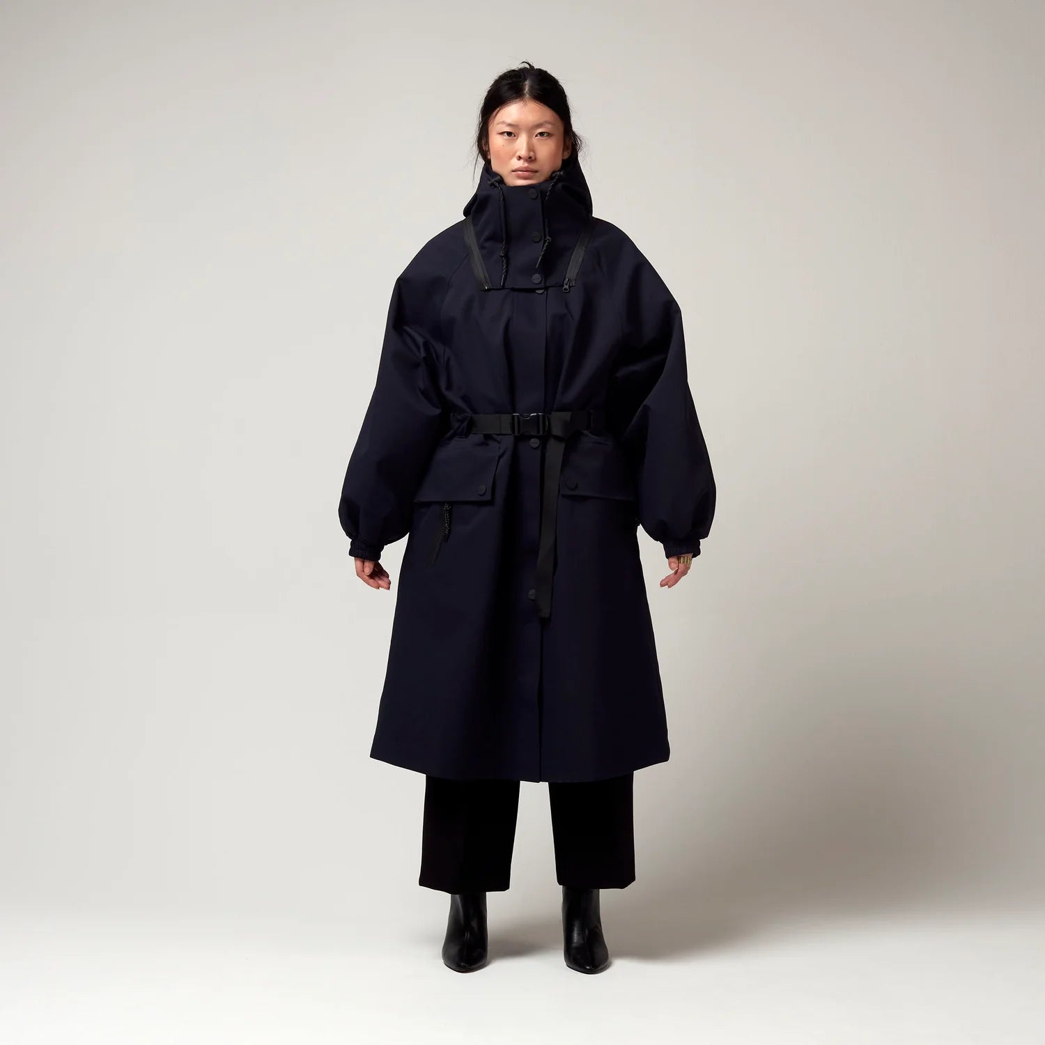 Dunkel Blauer Parker, mit Verstellbarer sowie abnehmbarer Kapuze, Taillierungs - Gurt um die Hüfte und Seiten Taschen. Model Bild, Ganzkörper Aufnahme.