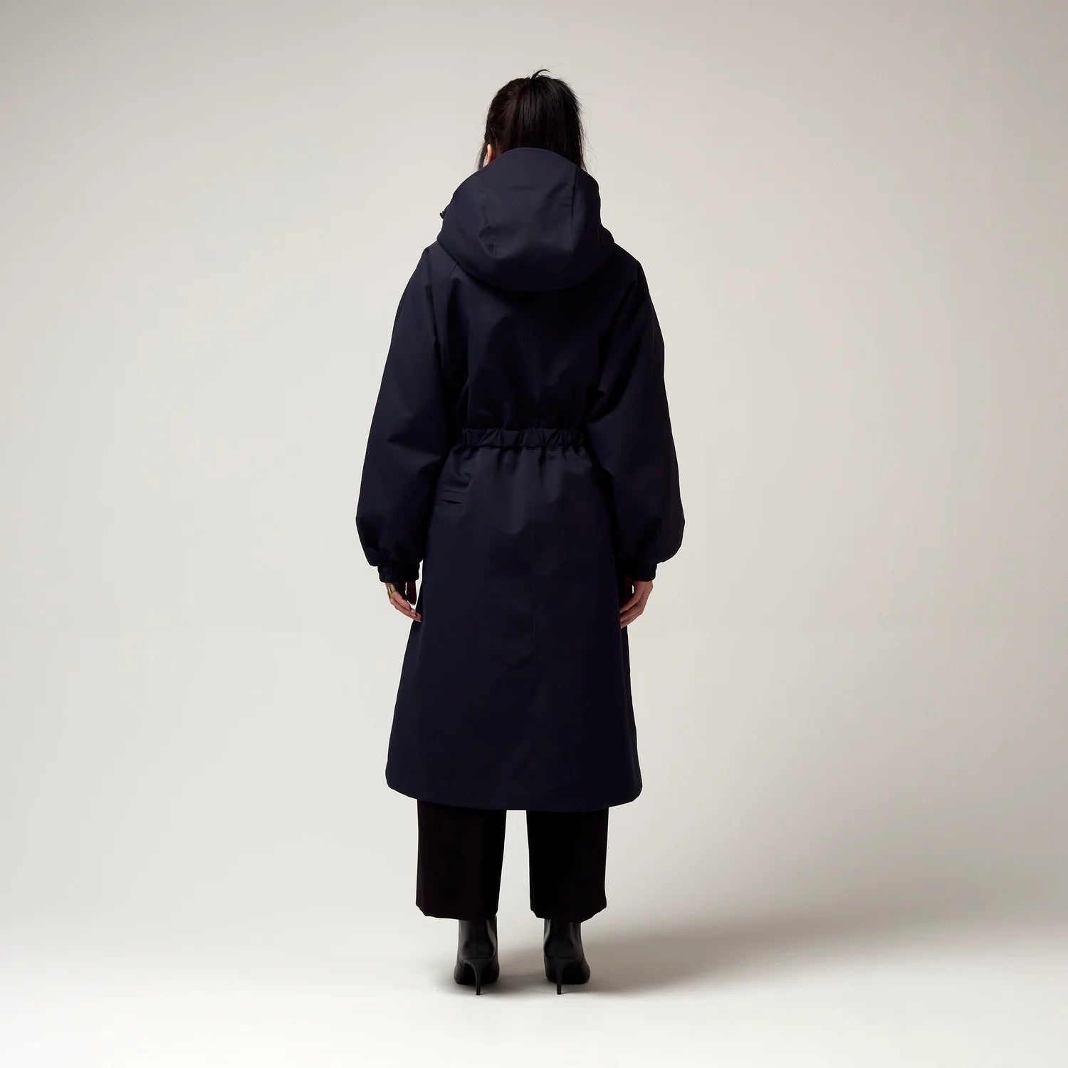 Dunkel Blauer Parker, mit Verstellbarer sowie abnehmbarer Kapuze, Taillierungs - Gurt um die Hüfte und Seiten Taschen. Model Bild, Ganzkörper Aufnahme, Rückseite.