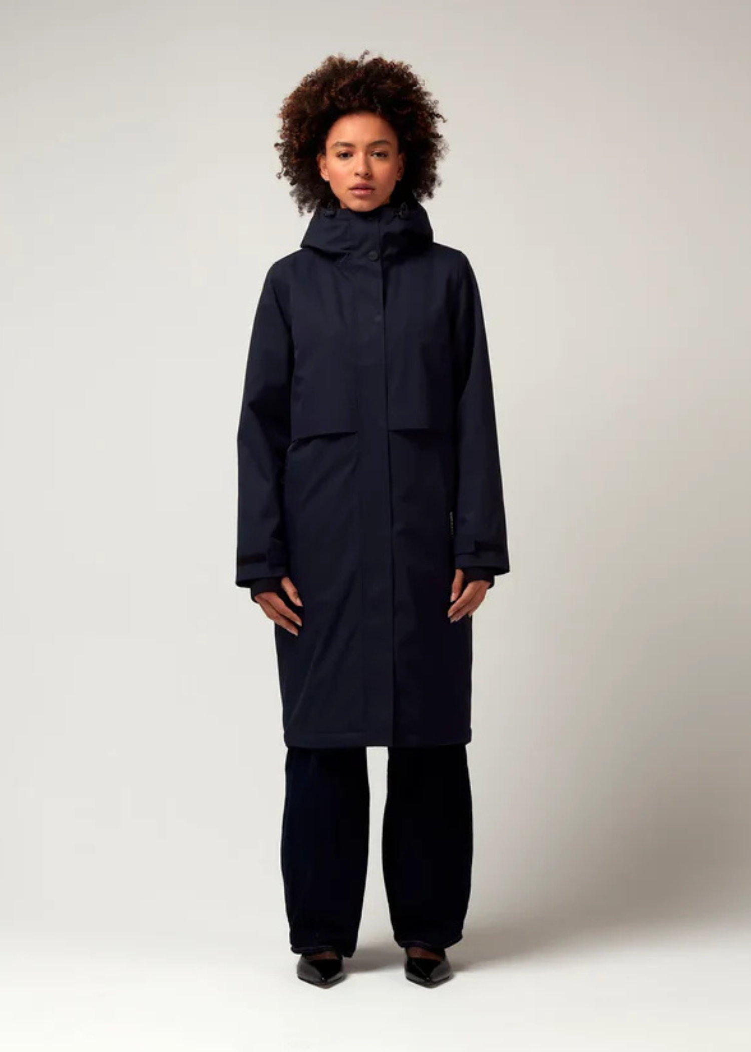 Dunkel Blauer Parker, mit verstellbarer Kapuze, Seitentaschen sowie Saum mit Daumen Loch. Glatte Oberfläche. Schwarze Hardware. Model Foto Ganzkörper Aufnahme.