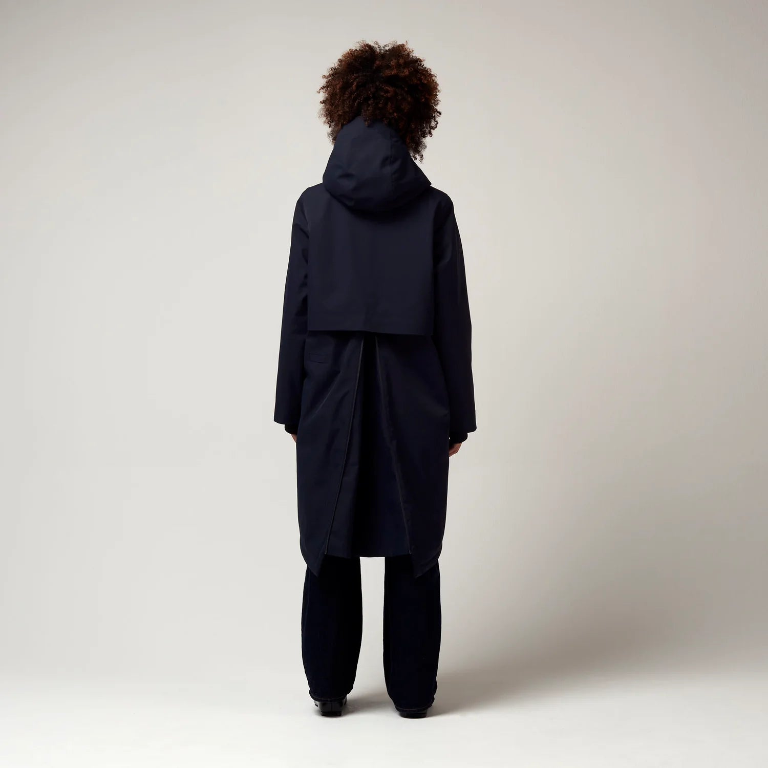 Dunkel Blauer Parker, mit verstellbarer Kapuze, Seitentaschen sowie Saum mit Daumen Loch. Glatte Oberfläche. Schwarze Hardware. Reißverschluss vom Saum der Jacke bis Hoch zum Oberen Rücken. Mit Integrierter Falte. Model Foto Ganzkörper Aufnahme, Rückseite. 