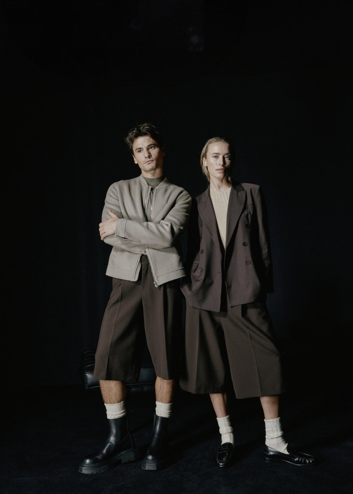 Ein männliches und ein weibliches Modell tragen die Bermuda Shorts vor einem schwarzen Hintergrund in der Ansicht von vorne. Die Shorts hat vorne Bundfalten.