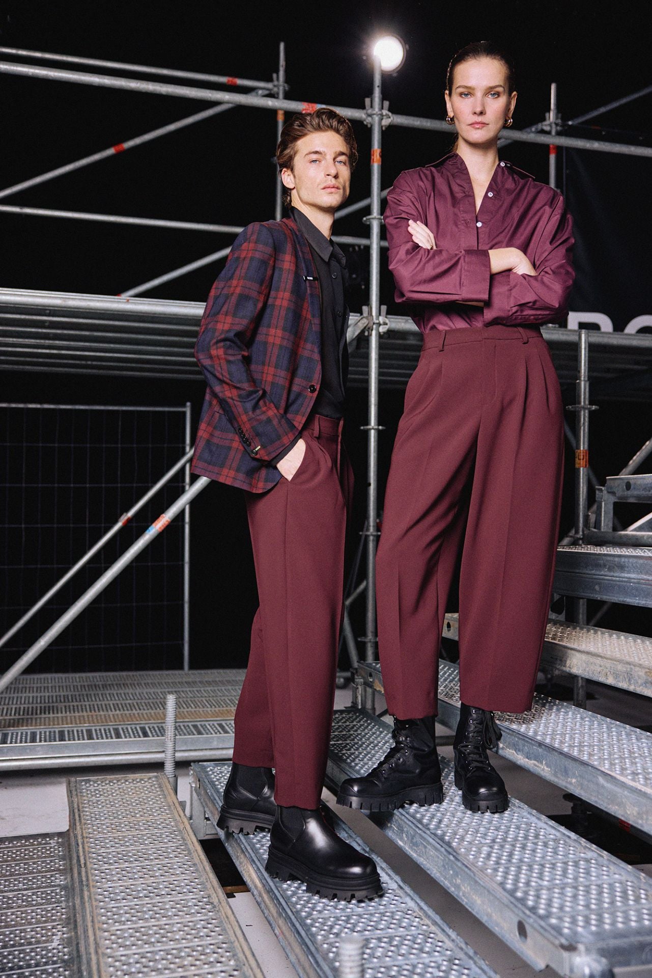 Anzugshose in Bordeaux mit Bundfalten und Mittelhohem Bund sowie Gürtelschlaufen. Seitlich integrierte Eingriffstaschen. Gerades Bein mit Verkürzter Schnitt. Getragen von jeweils einem Männlichen und einem Weiblichen Model. Crop Fit deutlich zu erkennen. Model Bild.